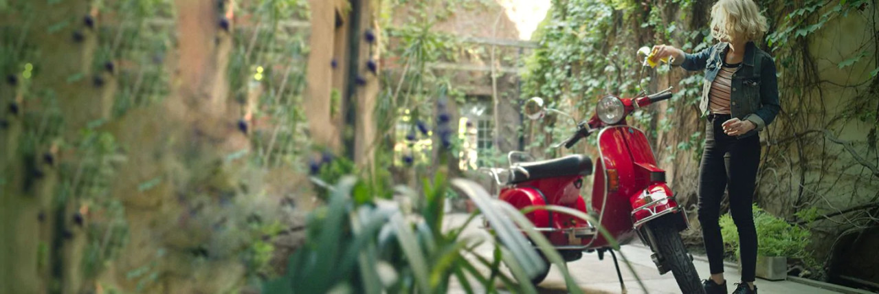 This image depicts a young woman smiling while cleaning her red vintage moped's wing mirror by the side of her house.