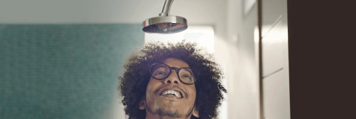 This image depicts the smiling face of a young man with glasses looking up at a shiny clean showerhead.