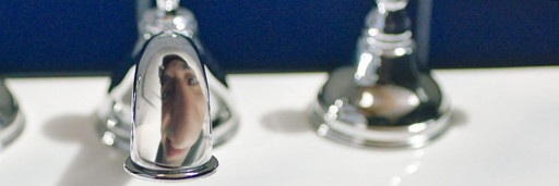 This image depicts the smiling reflection of a man's face in the spout of a tap. His face appears playfully distorted, exaggerating the size of his nose.